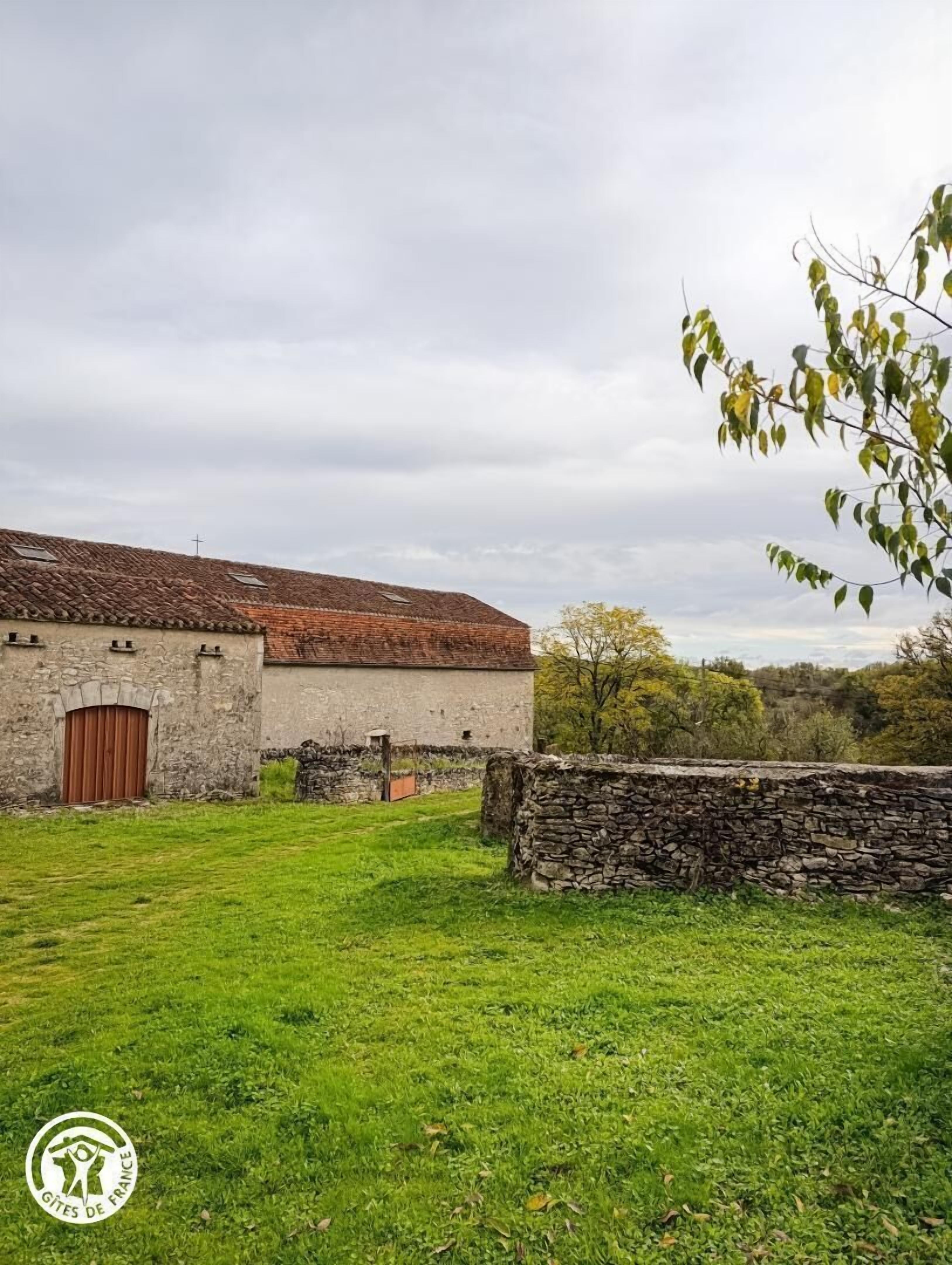 Un edificio rústico de piedra con techo de tejas rojas y puerta de madera, rodeado por un campo cubierto de hierba y un muro de piedra.