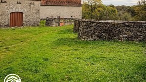 Um edifício rústico de pedra com telhado de telhas vermelhas e porta de madeira, rodeado por um campo gramado e um muro de pedra.
