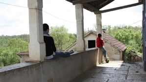 Varanda com duas pessoas sentadas, vista para paisagem rural com árvores e um edifício de pedra.