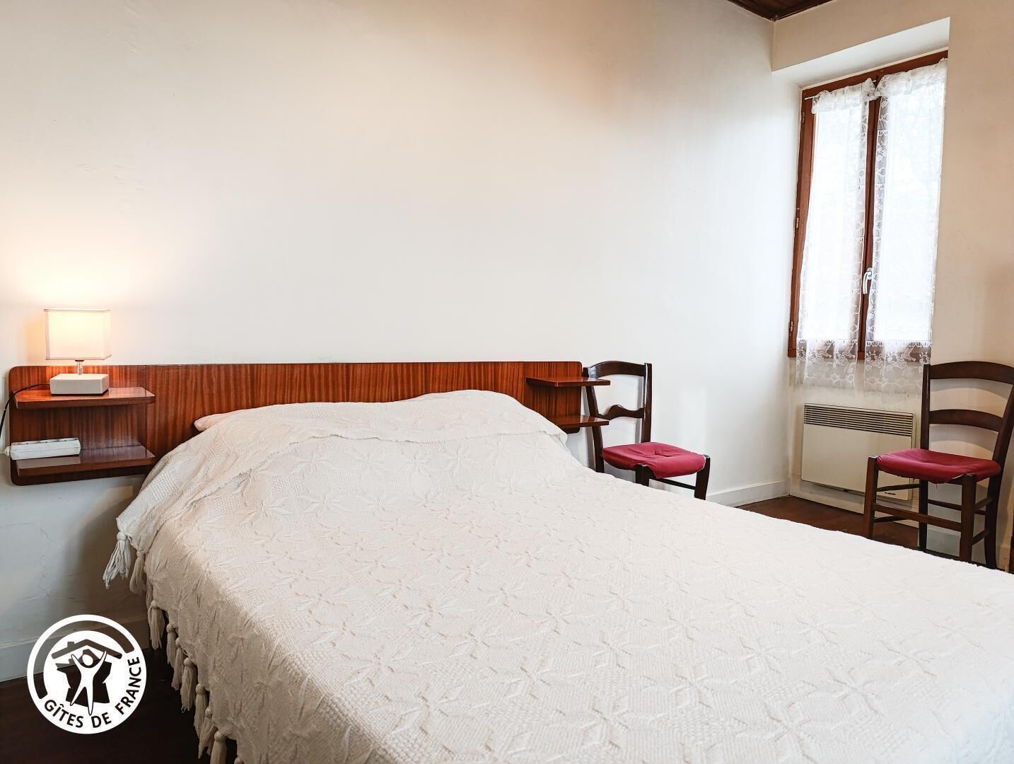 Un dormitorio con cama, mesita de noche, silla y ventana con cortinas.
