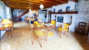A rustic interior with exposed wooden beams, a stone wall, a fireplace, wicker furniture, and a dining area with a staircase leading to an upper level. - Gîtes de France® - La Bergerie (LALBENQUE)