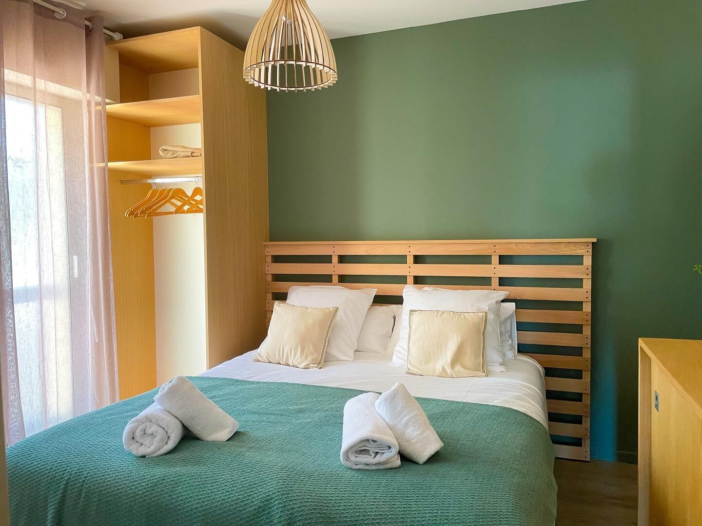 A neatly made bed with a green bedspread and two rolled towels, a wooden headboard, and a hanging light fixture.
