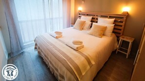 A neatly made bed with pillows and folded towels, a bedside table, and a window with sheer curtains. - Gîtes de France® - Le Loft Gramat (GRAMAT)