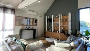 A spacious living area with a high ceiling, a built-in bookshelf, and a view of the outdoors. - Gîtes de France® - Le Loft Gramat (GRAMAT)