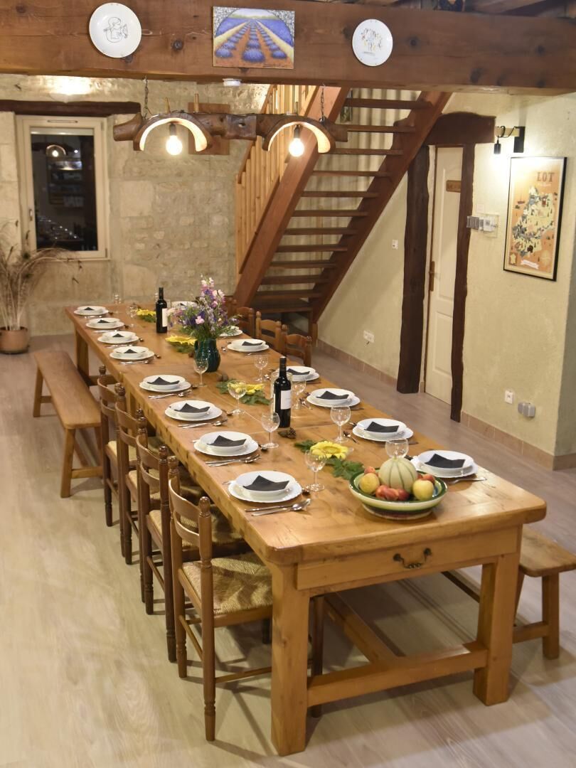 Una larga mesa de comedor de madera dispuesta con platos, vasos y un frutero, en una habitación con una escalera y vigas de madera.