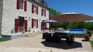 Edificio de piedra con contraventanas rojas, piscina, mobiliario exterior y una pérgola.