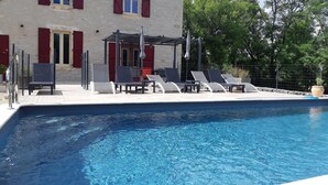 Una piscina con sillones de descanso y un edificio con contraventanas rojas al fondo.