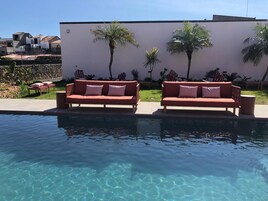 Un área de piscina con dos sofás rojos y una mesita, rodeada de palmeras y un muro blanco.
