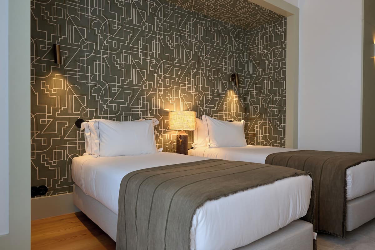 Hotellihuone, jossa on kaksi sänkyä, kuviollinen seinä ja yöpöytä, jossa on lamppu.