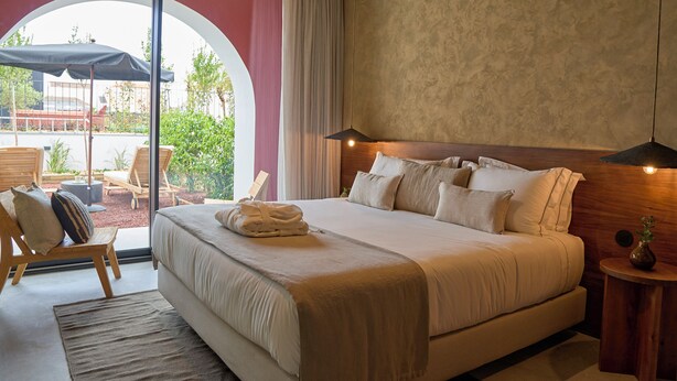 A modern hotel room with a large bed, a wooden headboard, a small side table, and a view of the outdoor area through a glass door. - The Farm - Azores Boutique House (Ponta Delgada)