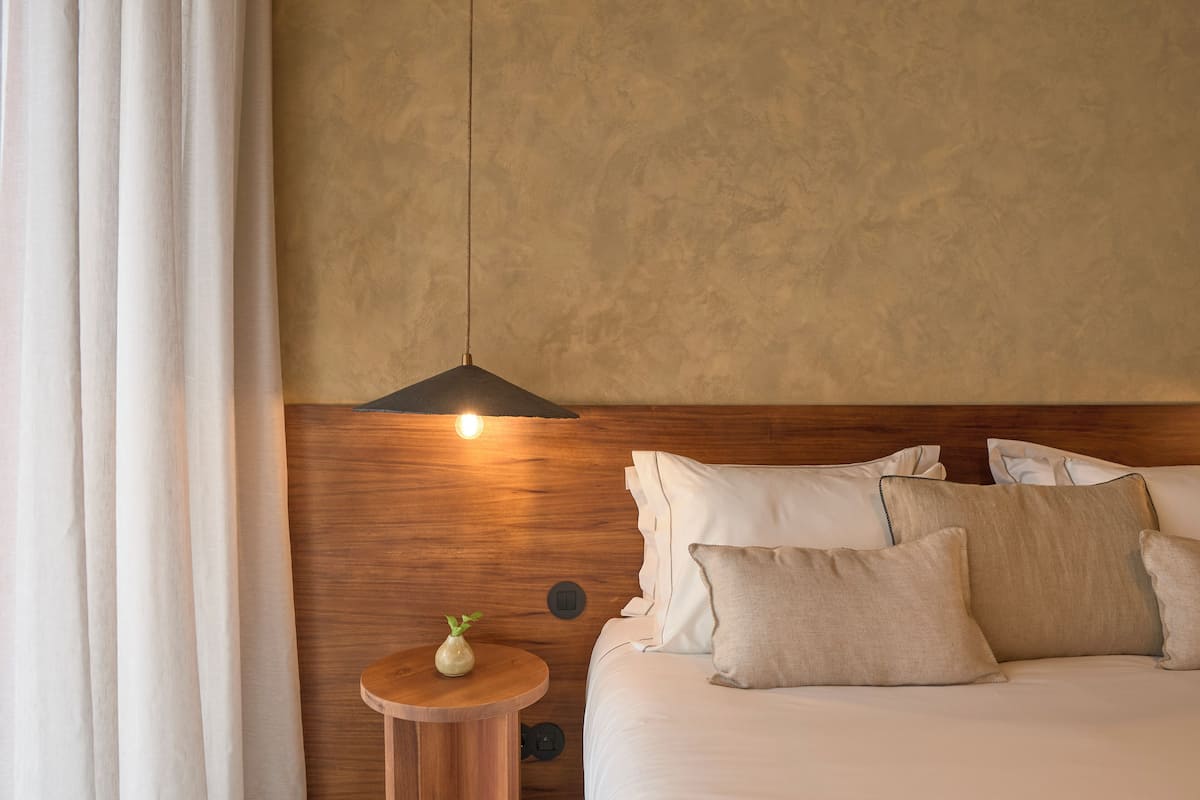 Hotellihuone, jossa on puinen sängynpääty, yöpöytä maljakolla ja useilla tyynyillä varustettu sänky.