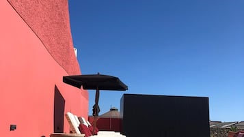A poolside area with lounge chairs and an umbrella, a red wall, and a clear blue sky.