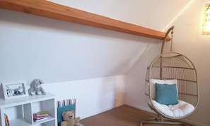 A room with a white wall, a wooden ceiling beam, a hanging chair with a cushion, a white shelf with books and a stuffed toy, and a framed picture.