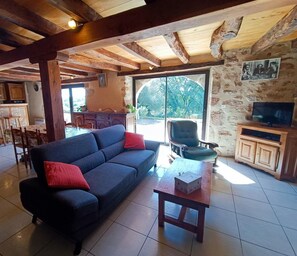 Ein großzügiger Wohnbereich mit einer Couch, einem Holztisch und einem Fernseher. Es gibt eine Küchenzeile mit einer Bar und einem Esstisch. Der Raum verfügt über eine Steinwand und ein großes Fenster.
