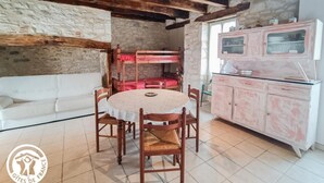 A rustic room with a stone wall, wooden beams, a bunk bed, a white sofa, a round wooden table with chairs, and a pink cabinet. - Gîtes de France® - L'Oustal (SAINT-SOZY)