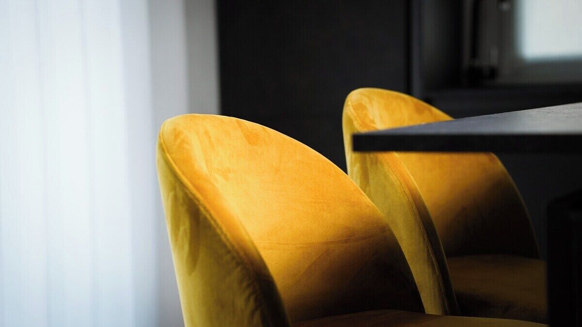 Dos sillas amarillas con una mesa oscura en una habitación con ventana y pared oscura.