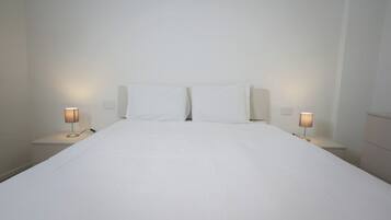 A neatly made bed with white linens in a simple room with two bedside tables, each with a lamp.