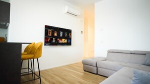 Un salon moderne avec un canapé gris, un fauteuil jaune et un téléviseur à écran plat fixé au mur, qui affiche une sélection de films.