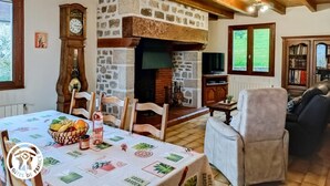 Un salon chaleureux avec une cheminée en pierre, des poutres apparentes, une table à manger agrémentée d’un panier de fruits et un canapé confortable.