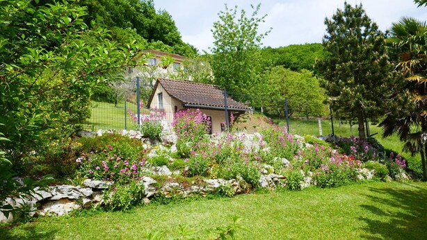 Une maison de campagne au toit recouvert de tuiles, entourée d’un jardin bien entretenu où poussent différentes espèces de plantes à fleurs, et bordée d’un mur en pierre.
