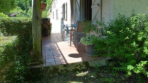 Un patio couvert avec une table et des chaises, entouré de verdure et avec une maison en arrière-plan.