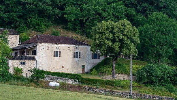 Une maison en pierre avec un toit de tuiles, entourée d’une végétation luxuriante et d’un mur de pierre.