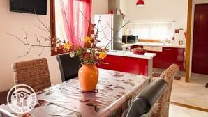 Ein Essbereich mit einem Tisch für zwei Personen, einer Blumenvase und Blick in eine Küche mit roten Schränken und einer Mikrowelle.