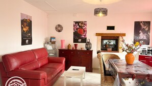 Ein Wohnzimmer mit rotem Ledersofa, einem weißen Couchtisch, einem Korbstuhl, einem Esstisch mit floralem Tischläufer, einem Kamin und Wandbildern.