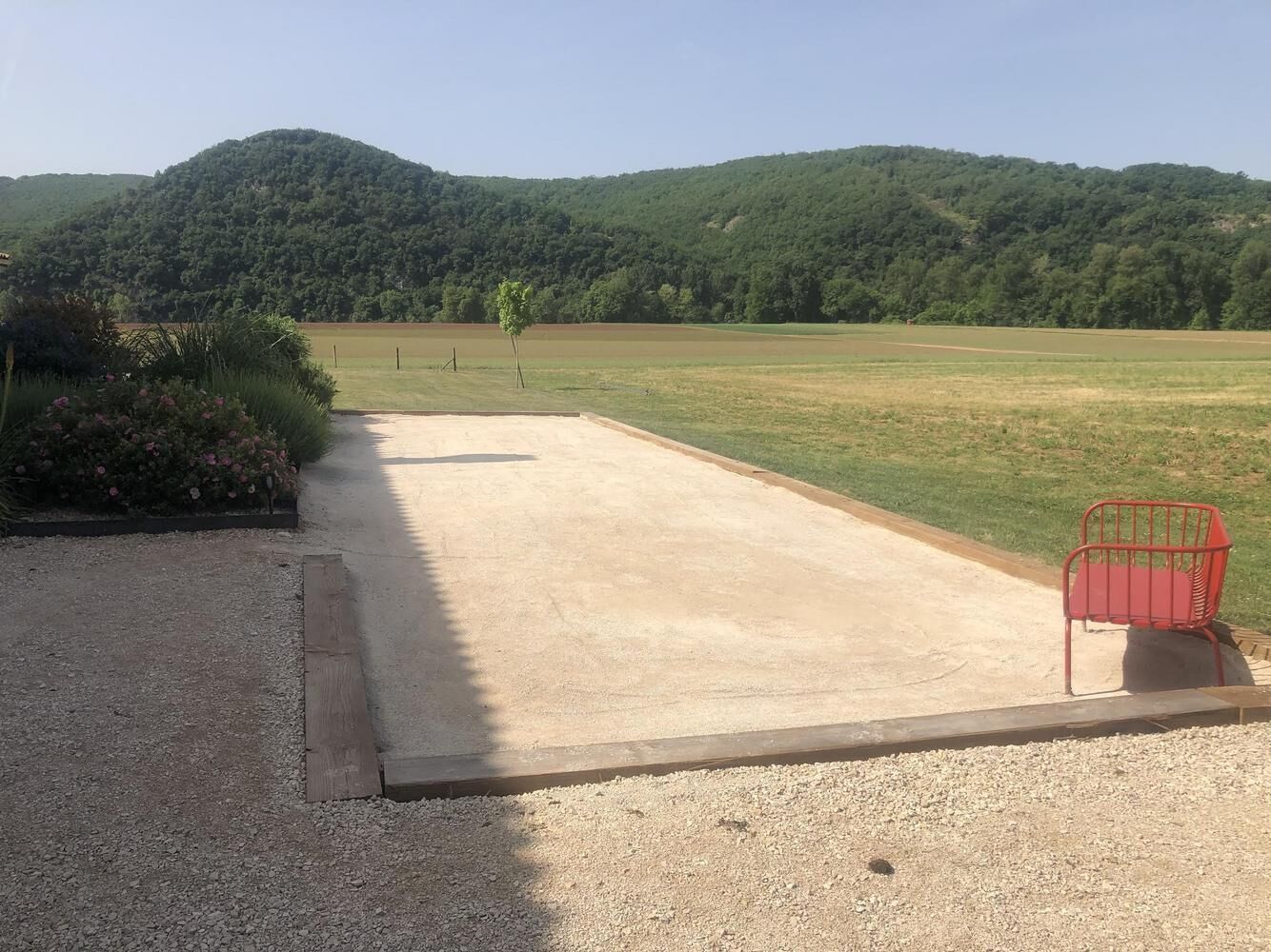 A paved area with a red metal bench, surrounded by gravel and a grassy field with a hill in the background.