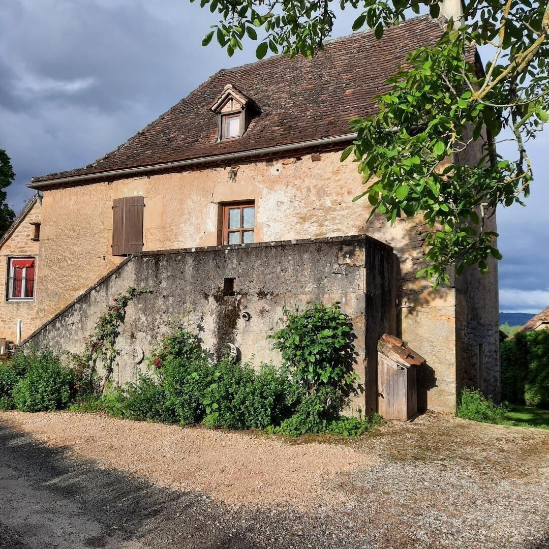 Un edificio rústico de piedra con techo inclinado, una pequeña ventana con contraventanas y un camino de entrada de grava.