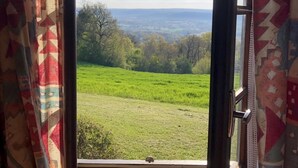 Een raam met uitzicht op een groen veld en bomen, gordijnen met een geometrisch patroon en een heldere lucht.