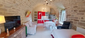 A cozy, stone-walled room with a TV, a sofa, a dining table, and a kitchen area. - Gîtes de France® - Le Pré aux Biches (ST MEDARD)
