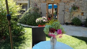 A stone building with a wooden door, a garden with potted plants, and a table with a vase of flowers.