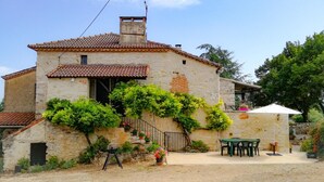 A stone house with a tiled roof, a small balcony, and a garden with potted plants and a table with chairs.