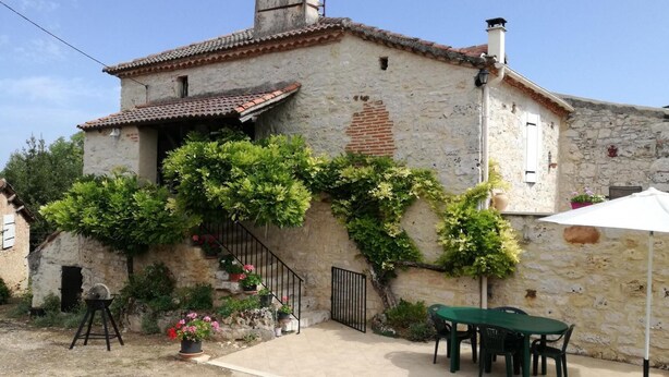 A rustic stone building with a tiled roof, a staircase with potted plants, and a table with chairs outside.
