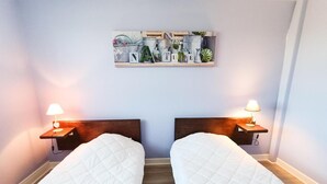 A room with two beds, each with a wooden headboard and a bedside table with a lamp. Above the beds, there is a wall-mounted shelf with various plants and gardening tools. - Gîtes de France® - L'Escurol (BRETENOUX)