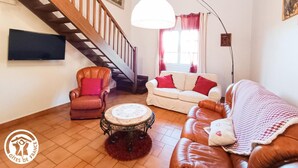 A living room with a wooden staircase, a sofa, a chair, a round table, and a TV mounted on the wall. - Gîtes de France® - L'Escurol (BRETENOUX)