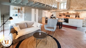 Uma sala de estar ampla com um sofá, uma mesa de centro redonda e uma mesa de jantar com cadeiras. Uma cozinha visível ao fundo apresenta uma parede de pedra e vigas de madeira.