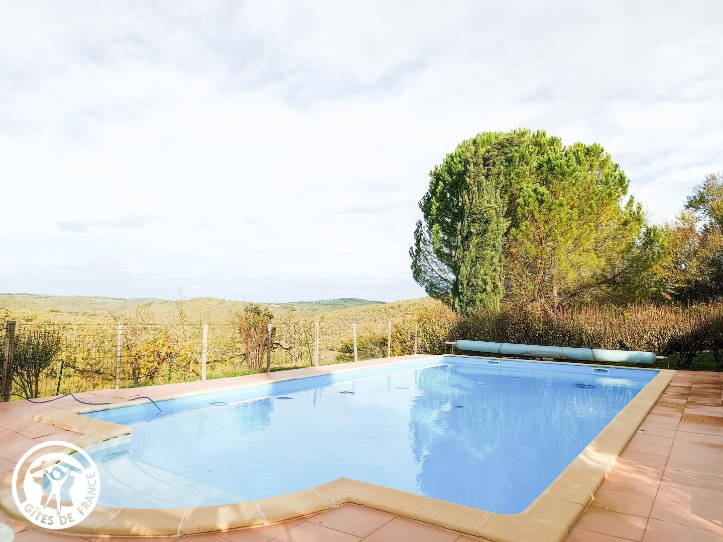 A rectangular swimming pool with a tiled deck, surrounded by a fence and trees, with a view of distant hills.
