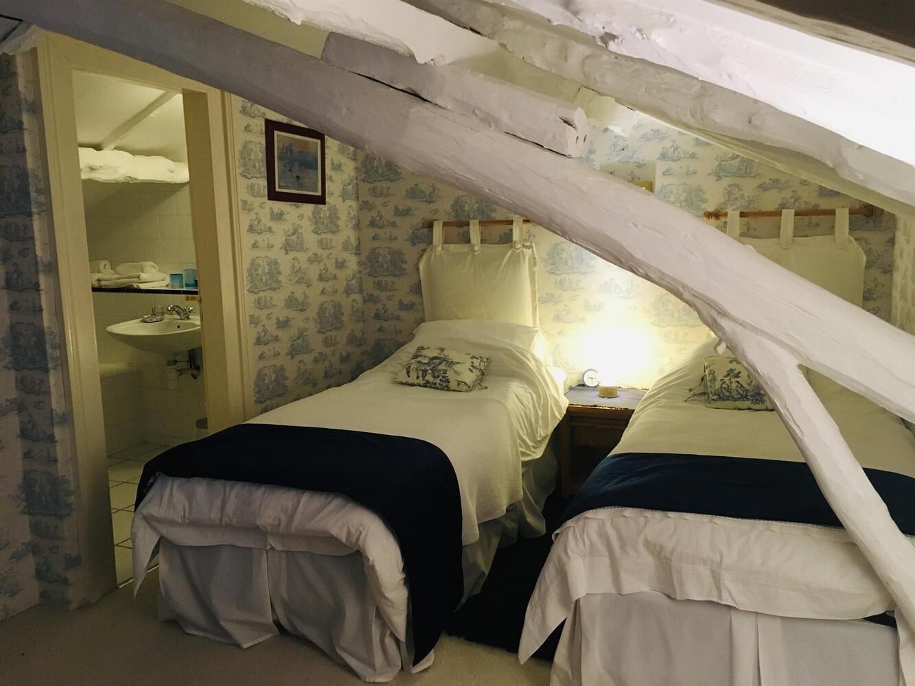 Un dormitorio con dos camas individuales, una mesita de noche con lámpara y un baño con lavabo y espejo.