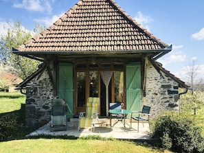 A stone cottage with a tiled roof, green shutters, and a small patio with outdoor furniture.