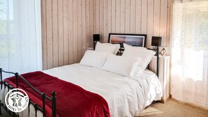 Un dormitorio con pared revestida de madera, cama con ropa de cama blanca y una manta roja, cabecero de metal negro, lámpara colgante y mesita de noche con una lámpara y un pequeño objeto decorativo.