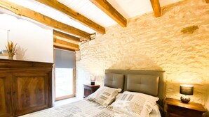 A bedroom with a stone wall, a wooden ceiling, a bed with a patterned bedspread, two bedside tables with lamps, and a door leading to a balcony.
