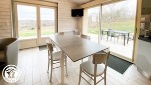 A dining area with a large table and chairs, a television mounted on the wall, and large sliding glass doors leading to an outdoor patio with a view of the countryside.
