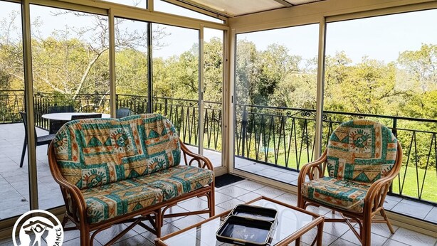 A covered patio with wicker furniture and a glass table, overlooking a lush garden. - Gîtes de France® - La Cigale (SAINT PROJET)
