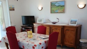 A dining area with a table set for four, surrounded by red chairs. A chandelier hangs above the table, and there are wall-mounted light fixtures. A television is on a stand to the left, and a cabinet with a picture frame is to the right. - Gîtes de France® - La Cigale (SAINT PROJET)
