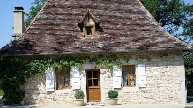 A stone house with a pitched roof, a wooden door, and shutters. There are two potted plants in front.