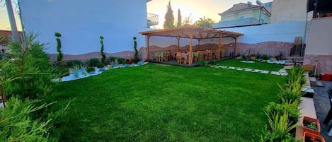 A well-maintained lawn with a pergola and seating area, surrounded by potted plants and a clear sky.