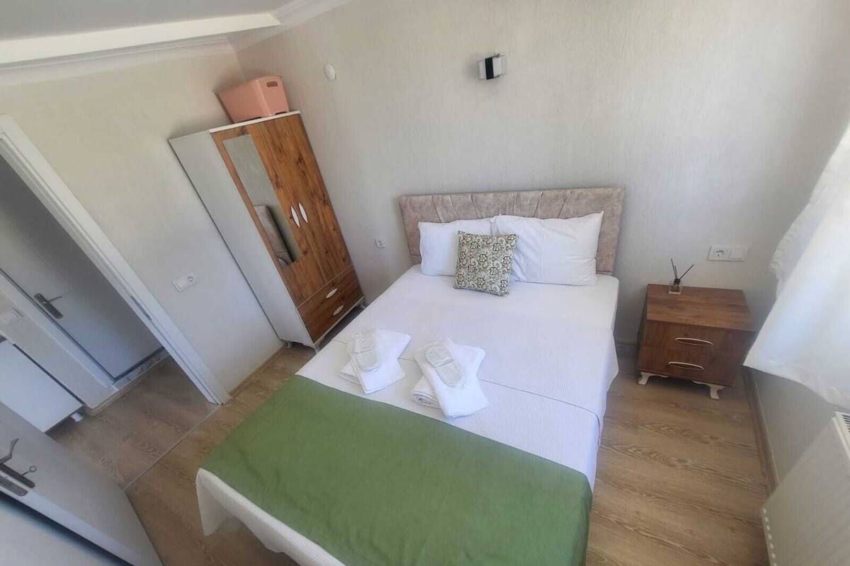 A neatly arranged bedroom with a bed, a wooden wardrobe, a small bedside table, and a green bedspread.