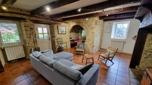 A cozy living room with a grey sofa, wooden chairs, a stone wall, and a tiled floor. - Gîtes de France® - Gîte de Larigaldie (SAINT CIRGUES)